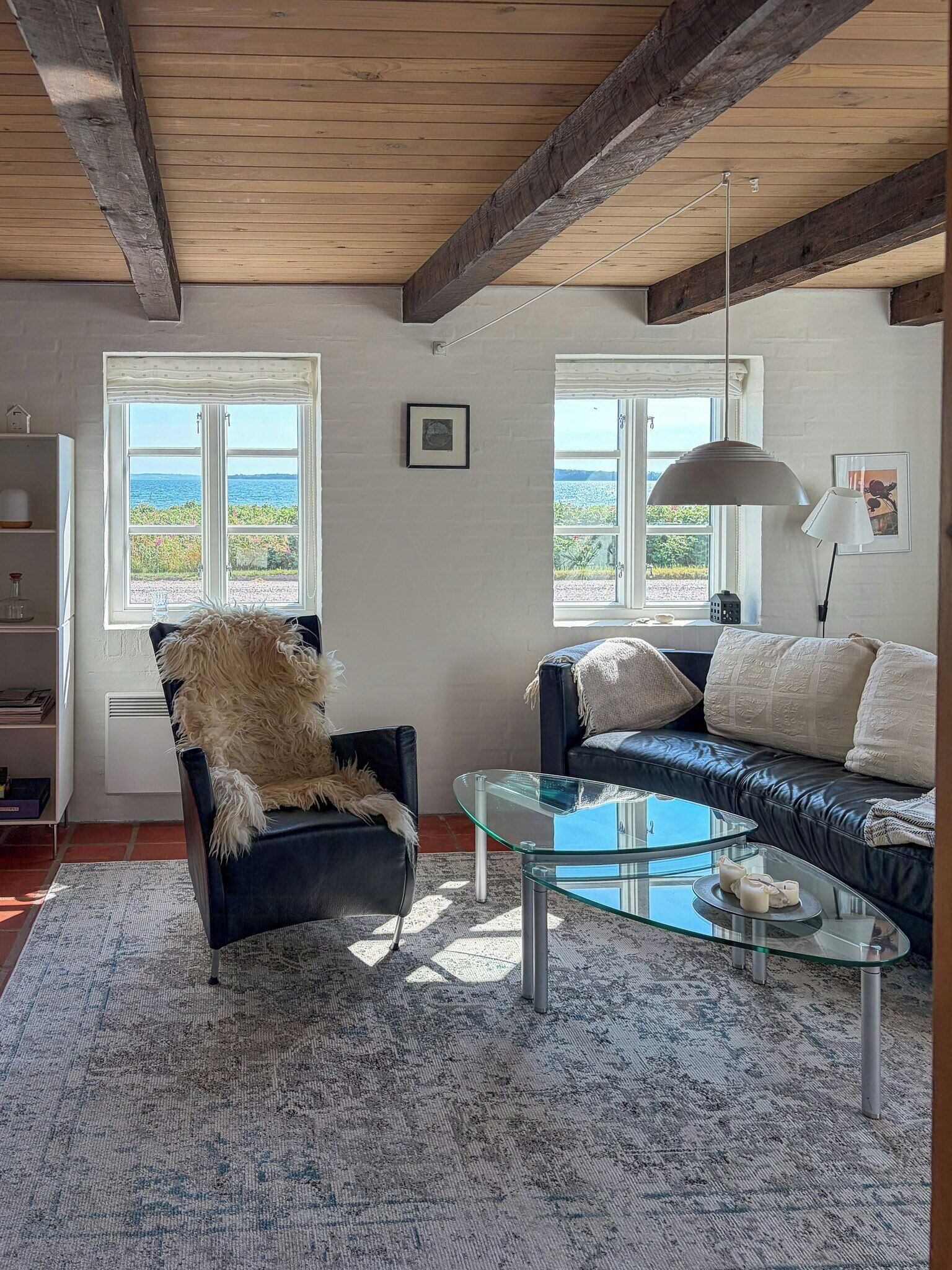 A living room with a glass coffee table, a black leather sofa, a fluffy chair, and a view of the sea through large windows.