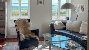 A living room with a glass coffee table, a black leather sofa, a fluffy chair, and a view of the sea through large windows.
