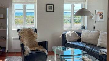 A living room with a glass coffee table, a black leather sofa, a fluffy chair, and a view of the sea through large windows.