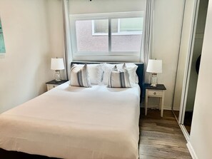 Ein ordentlich bezogenes Bett mit weißer Bettwäsche und gestreiften Kissen, zwei Nachttische mit Lampen, ein Fenster mit Jalousien und ein Holzboden.