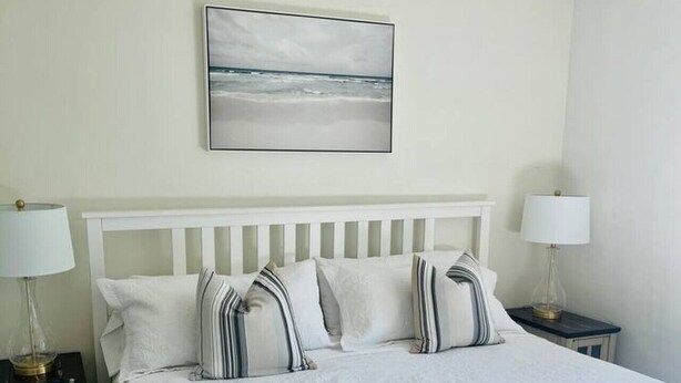Een slaapkamer met een wit bed, gestreepte kussens, een nachtkastje met een lamp en een schilderij aan de muur.