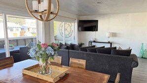 Een moderne woonkamer met een eethoek, een grijze zithoek, een houten eettafel met stoelen, een vaas met bloemen en een flatscreen tv aan de muur.