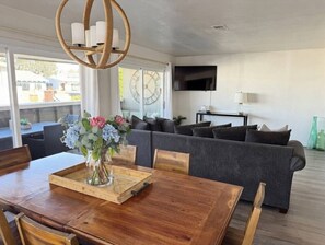 Una sala moderna con comedor, un sofá gris, una mesa de comedor de madera con sillas, un jarrón con flores y un televisor de pantalla plana montado en la pared.