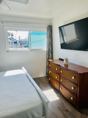 Ein Schlafzimmer mit Bett, Kommode, Fernseher und Fenster mit Blick auf Gebäude.