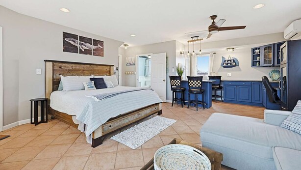 A bedroom with a large bed, a small table, and a chair. There is a kitchenette with a bar and chairs. - Spacious 5-bedroom WAVES OF HAMPTON I & II Combined (Hampton)