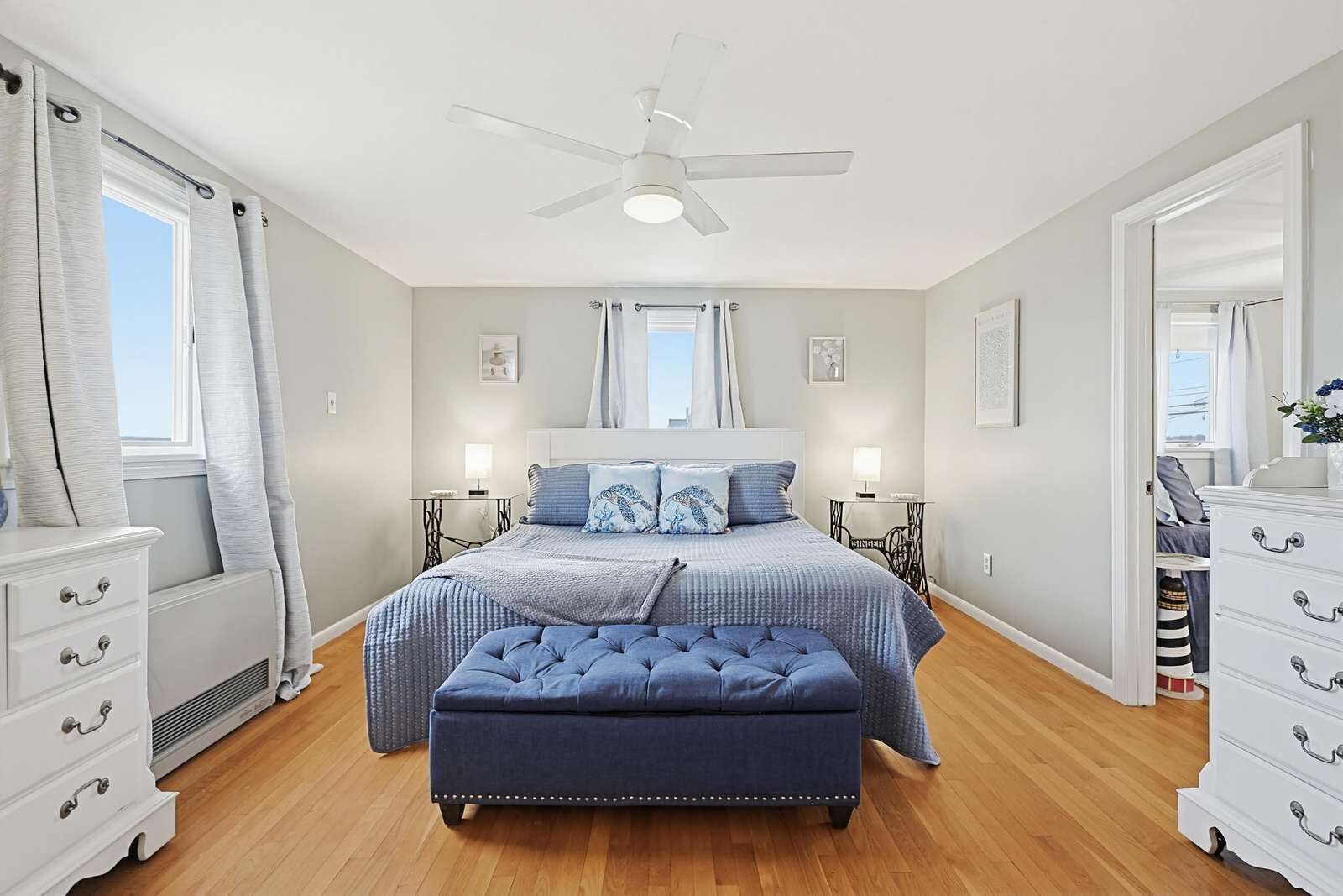 A bedroom with a bed, a ceiling fan, bedside tables, a dresser, and a window with curtains.
