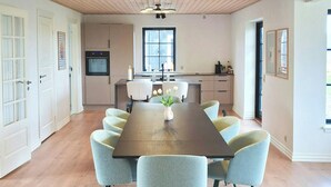 A dining area with a wooden table and six chairs, a kitchen with a stove and cabinets, and a large window. - Newly renovated holiday home near HimmerLand (Farsø)