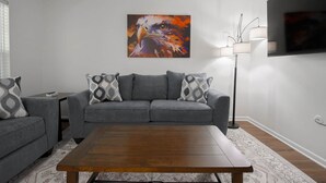 Un salon avec un canapé gris, des coussins à motifs, une table basse en bois, un lampadaire et un tableau accroché au mur.