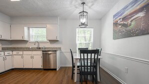 Une cuisine avec des meubles blancs, un lave-vaisselle en acier inoxydable, une fenêtre avec des stores, une table à manger avec des chaises et un luminaire au plafond.