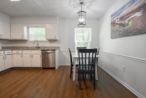 A kitchen with white cabinets, a stainless steel dishwasher, a window with blinds, a dining table with chairs, and a ceiling light fixture. - Eagles Landing-easy access to AU (Auburn)