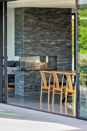 A modern interior with a stone fireplace, a glass door leading to a balcony, and a seating area with wooden chairs. - Summer house with sea view high in Handrup (Ebeltoft)