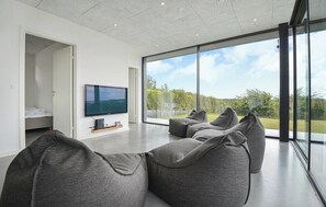 A modern living room with a large TV, grey sofas, and a view of greenery through large windows. - Summer house with sea view high in Handrup (Ebeltoft)