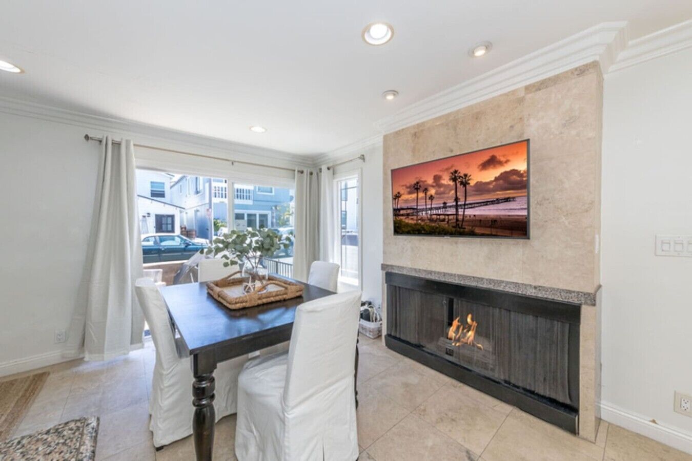 A dining area with a fireplace, a painting of a sunset, and a view of the street outside.
