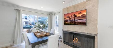 A dining area with a fireplace, a painting of a sunset, and a view of the street outside.