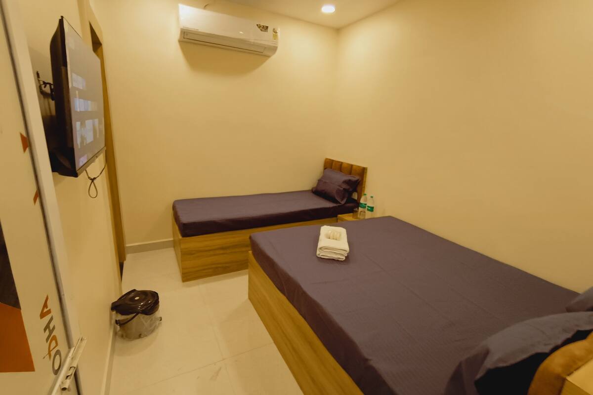 A small room with two single beds, a wall-mounted air conditioner, a TV, and a trash bin.