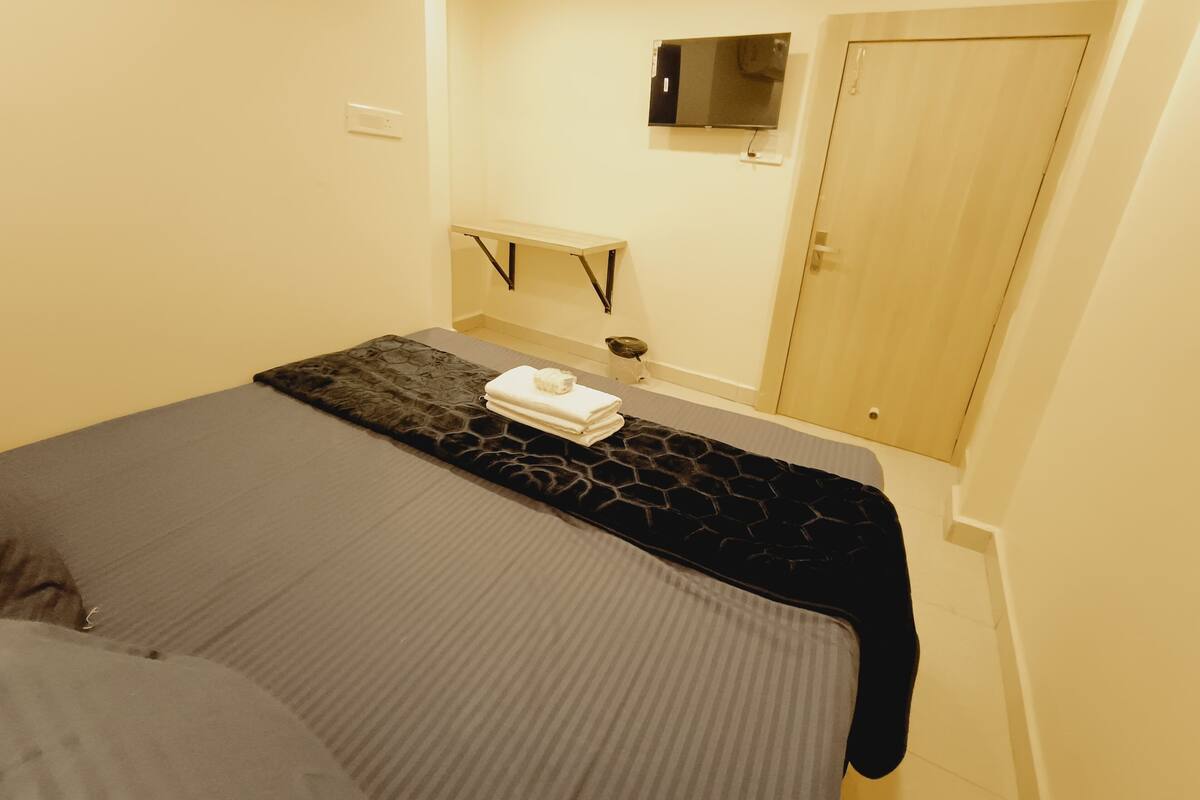 A hotel room with a bed, a bedside table, a TV mounted on the wall, and a door.