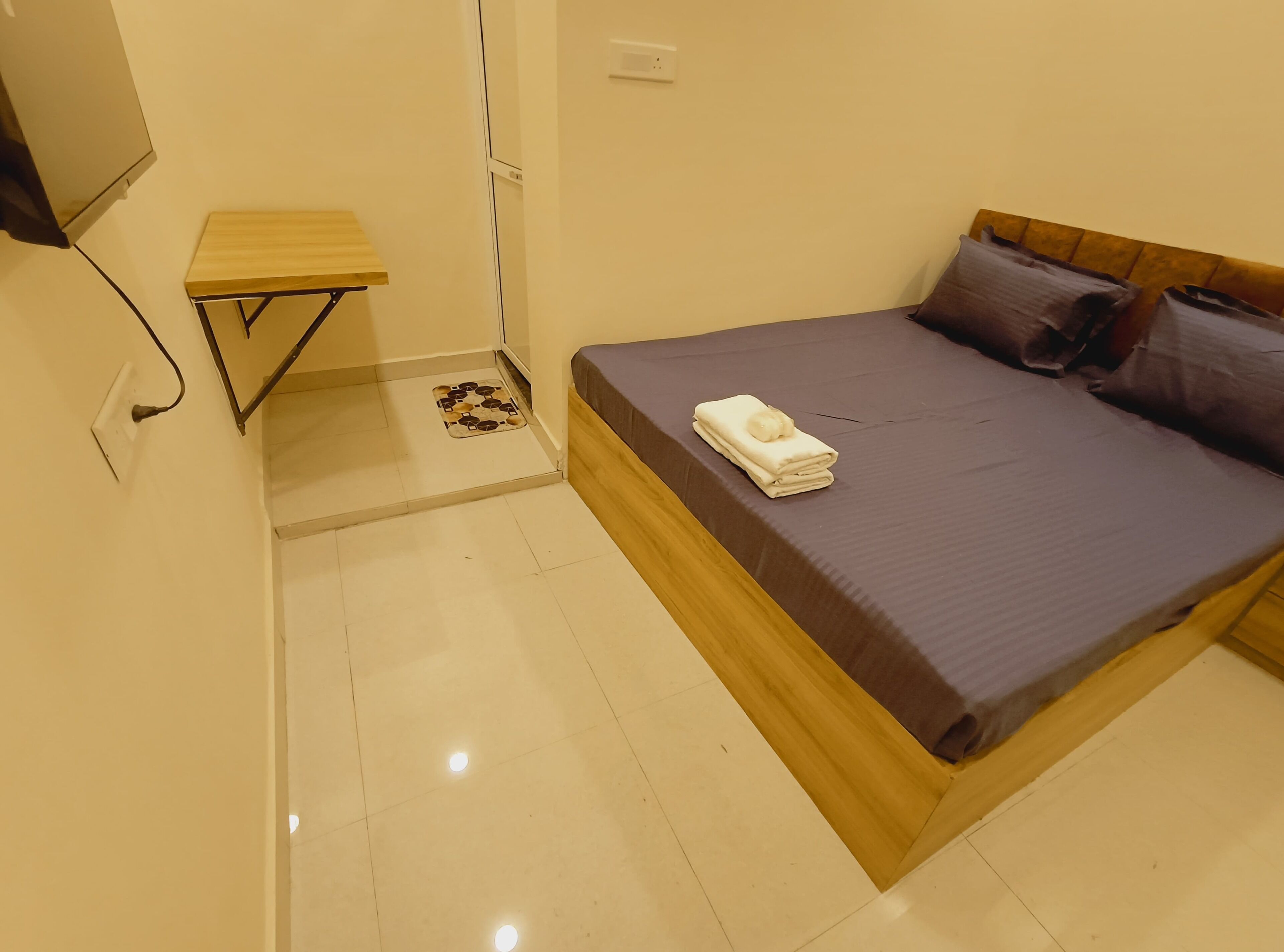 Un dormitorio con cama, una mesita de madera, televisor montado en la pared y un enchufe.