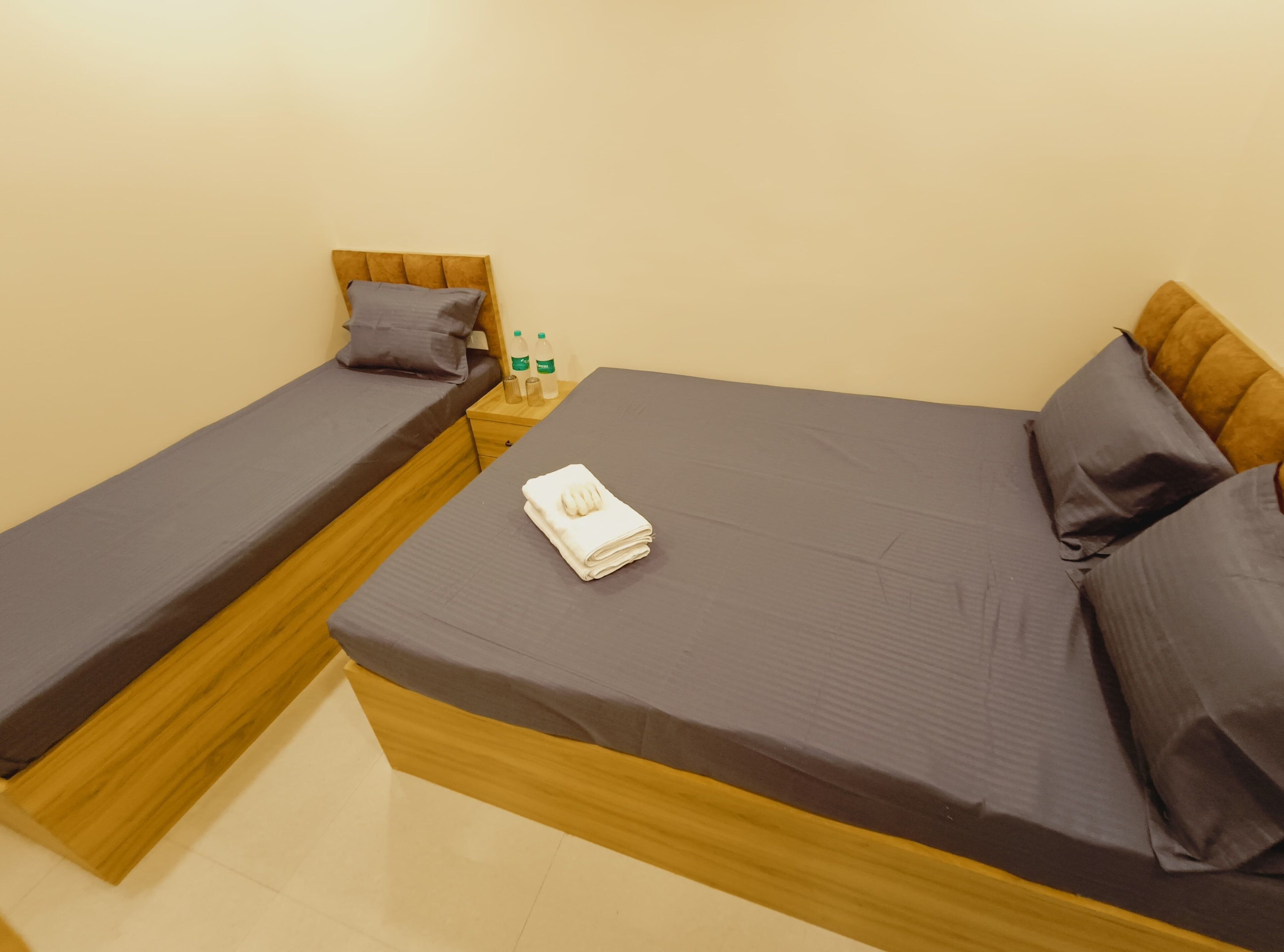 Una cama de matrimonio con estructura de madera, una cama individual con estructura de madera, una mesita de noche con una botella y un vaso, y una toalla doblada sobre la cama individual.