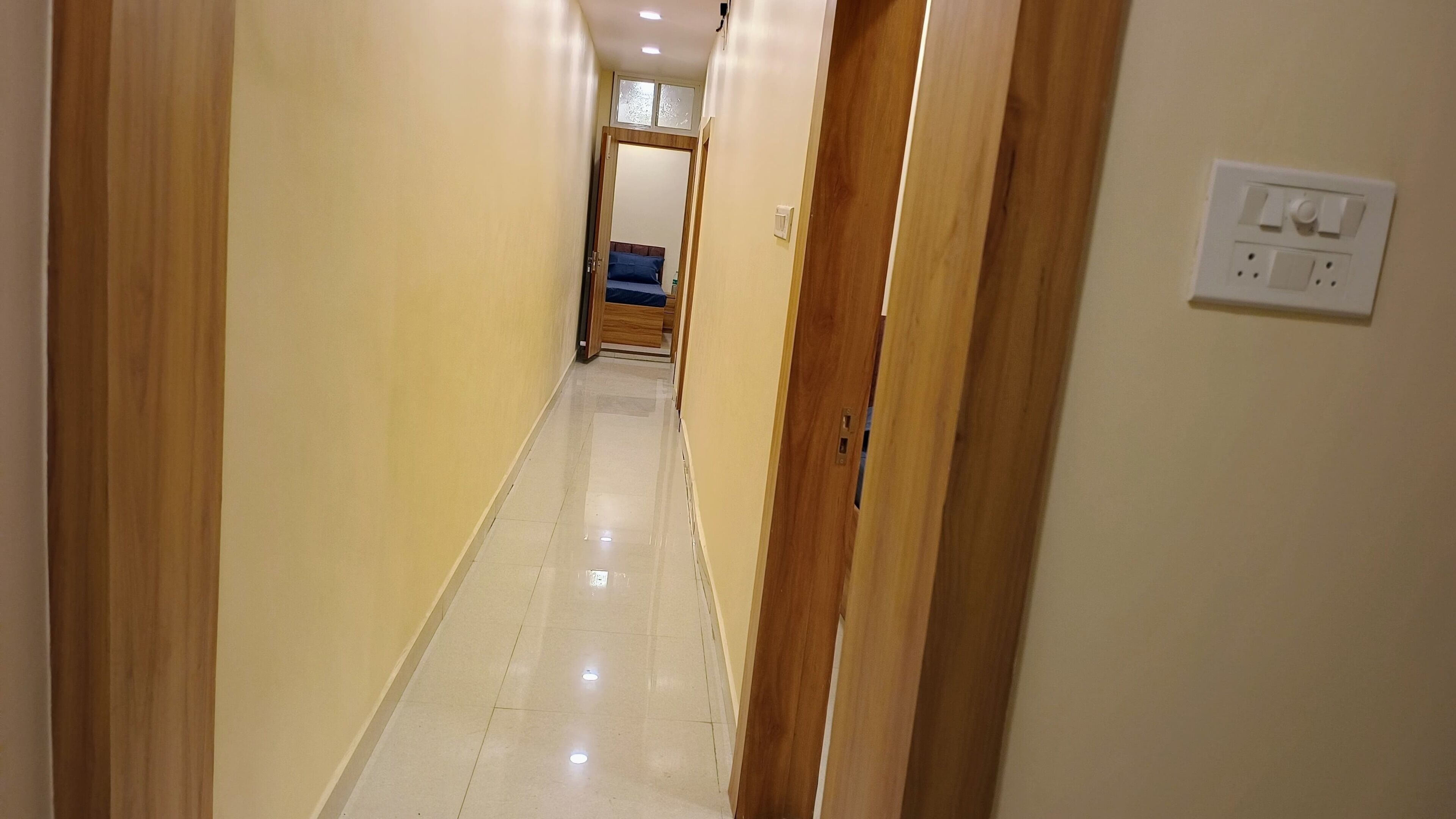 A narrow hallway with a wooden door on the right, a light switch on the wall, and a bed with a blue cover in the distance.