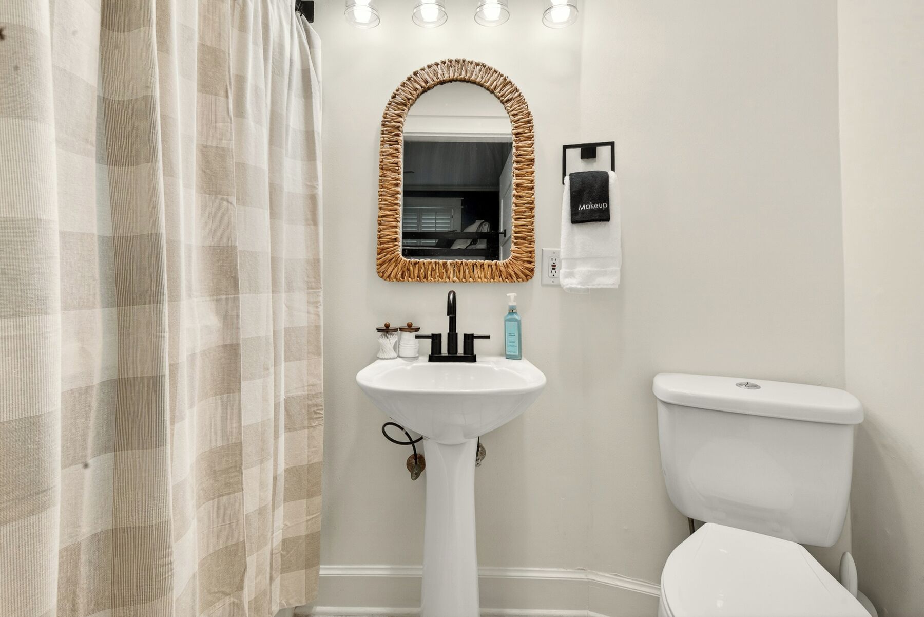A bathroom with a white sink, a toilet, a mirror with a woven frame, and a towel rack with a 'Makeup' sign.