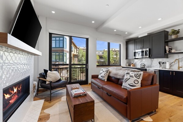 A modern living room with a fireplace, a leather sofa, and a dining area with dark cabinets and a kitchen. - Upscale Riverfront Condo- BC Gondola, Dog Friendly (Avon)