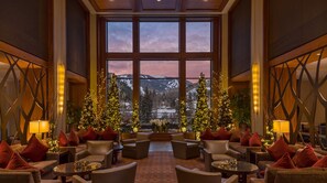 A spacious lobby with multiple seating areas, large windows offering a mountain view, and Christmas trees adorned with lights. - Upscale Riverfront Condo- BC Gondola, Dog Friendly (Avon)