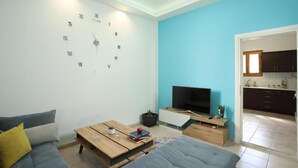 A modern living room with a large wall clock, a flat-screen TV, and a kitchen visible through an open doorway.