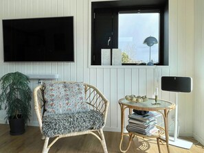 Ein Zimmer mit an der Wand montiertem Fernseher, einem Korbstuhl mit Blumenkissen, einem kleinen runden Tisch mit Lampe und einer Topfpflanze.