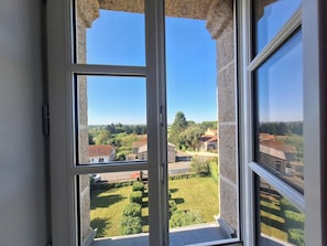 Blick durch ein Fenster auf eine steine Mauer, einen klaren Himmel und eine grüne Landschaft mit Gebäuden.