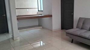 Een moderne woonkamer met een grijze bank, een klein keukenhoekje met een aanrecht, en een betegelde vloer.