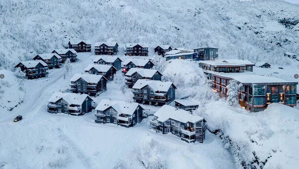 Un complejo turístico nevado con varias construcciones de estilo chalet, un telesilla y montañas al fondo.