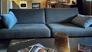 Una sala moderna con un sofá, una mesita de centro con revistas, una lámpara y vista a la cocina.