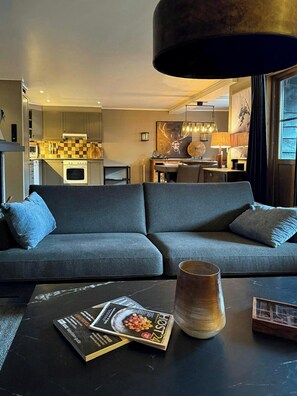Ein modernes Wohnzimmer mit einer Couch, einem Couchtisch mit Zeitschriften, einer Lampe und Blick in die Küche.