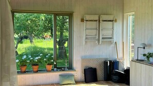 Een kamer met houten wanden, een schuin dak, een raam met uitzicht op groen, een bank met potplanten en een kleine tafel met een lamp.