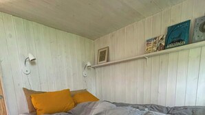 Een bed met een grijs-witte deken, twee mosterdgele kussens en een plank met boeken en een ingelijst foto.