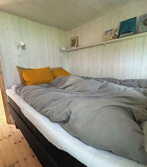 Un lit avec une couette grise et blanche, deux coussins couleur moutarde et une étagÚre avec des livres et un tableau encadré.