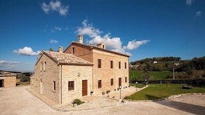 A stone building with a tiled roof, a paved courtyard, and a grassy area with a fence. - Historic villa with pool in Marche hills (Sant'Angelo in Pontano)