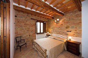 Un dormitorio con pared de piedra, una cama, una silla, una mesita de noche y una ventana.