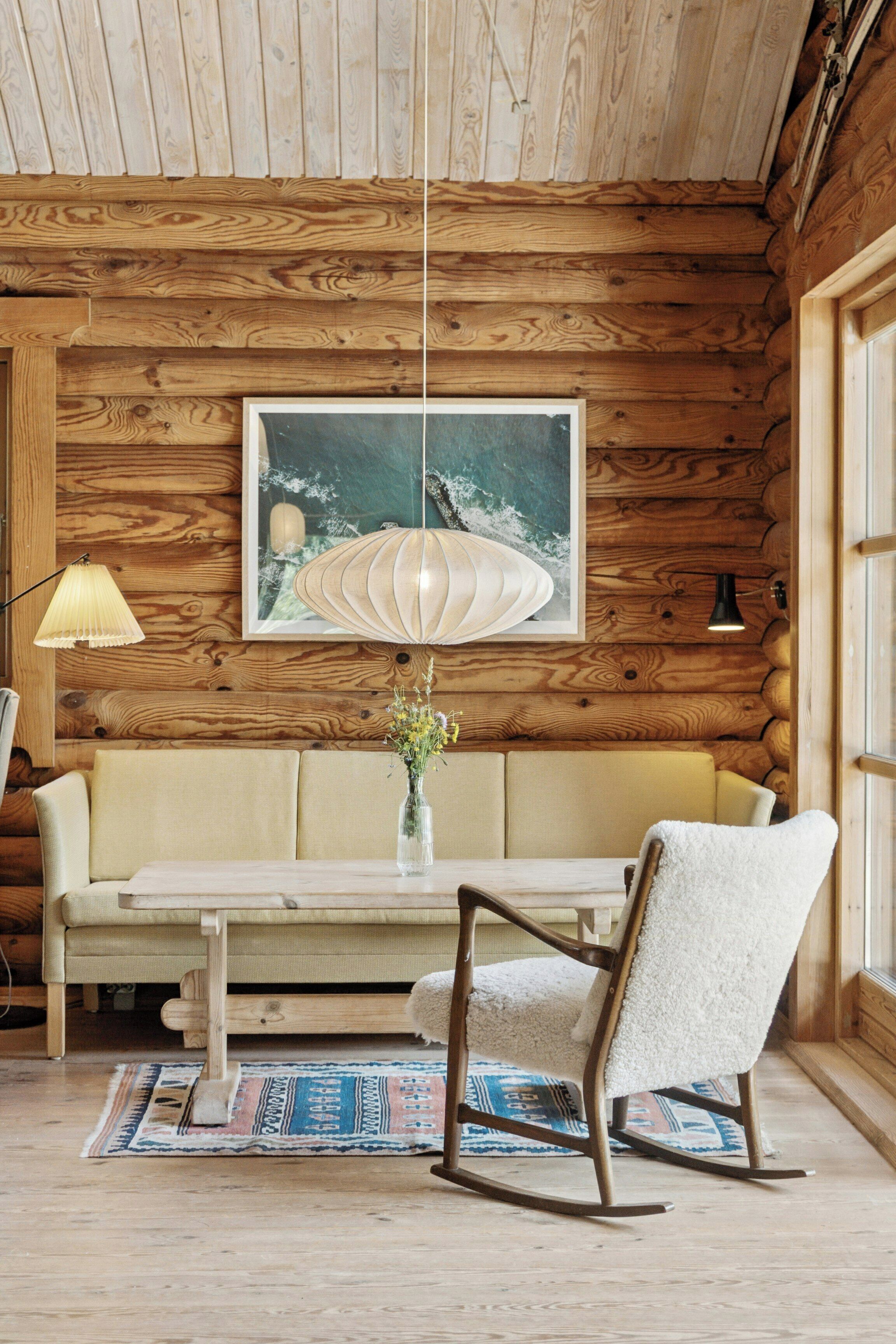 A log cabin interior with a wooden ceiling, a framed artwork, a sofa, a rocking chair, a table, and a lamp.