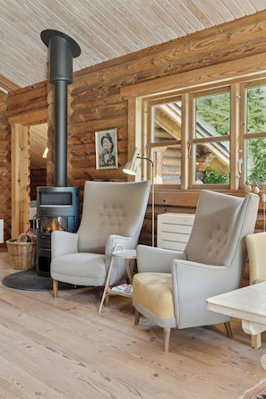 Ein rustikales Holzhaus-Interieur mit Kaminofen, zwei Polsterstühlen, einer Stehlampe und einem Fenster mit Blick auf Grünflächen.