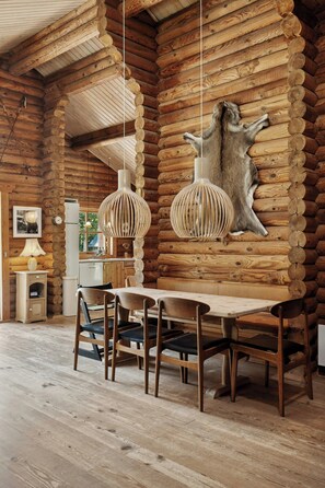 Ein rustikaler Innenraum einer Blockhütte mit Esstisch, Stühlen, Pendelleuchten und einem aufgehängten Tierkopf an der Wand.