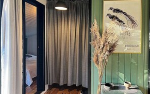Ein Zimmer mit einem Bett, einem Vorhang, einer Hängelampe, einem Rahmenbild mit Vögeln und einer Vase mit Trockenpflanzen.