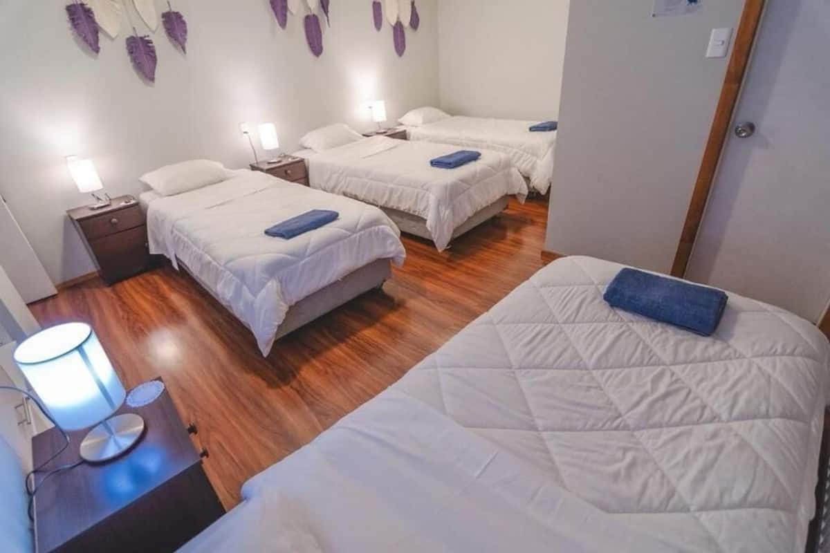 A hotel room with three beds, a nightstand with a lamp, and a wooden floor.