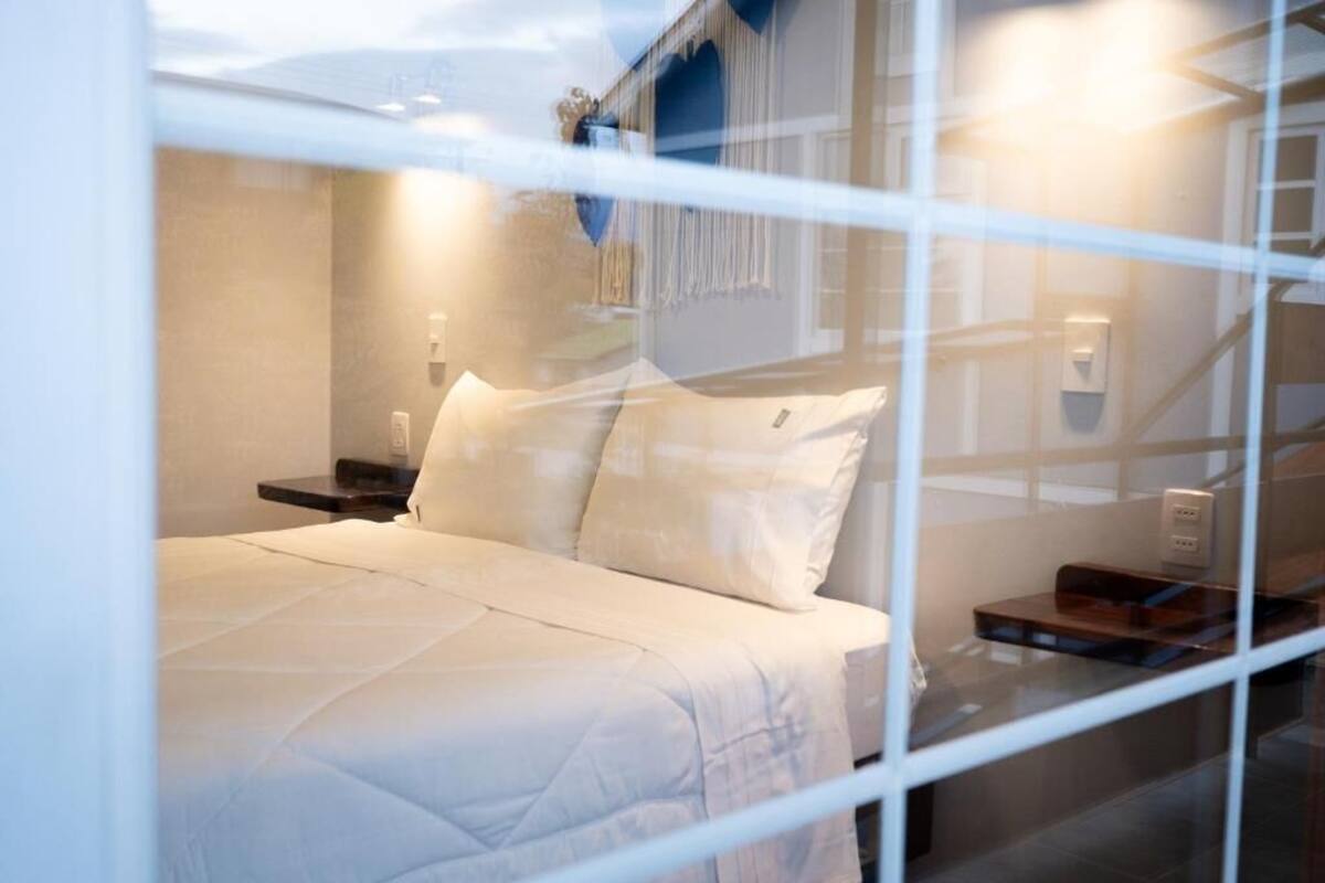 A bed with white bedding, a nightstand, a wall-mounted light, and a window with a view of a building.