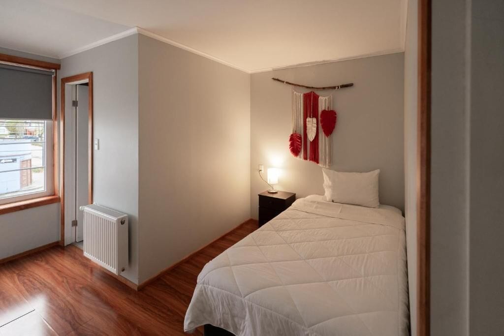 A bedroom with a bed, a nightstand, a wall-mounted heart decoration, a radiator, and a window.