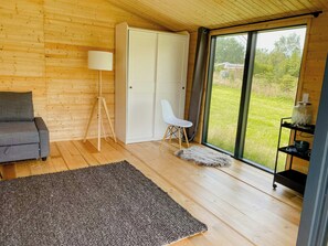 A room with a wooden floor, a grey sofa, a white chair, a lamp, a fur rug, and a sliding glass door leading to a grassy area. - Charming holiday home near the beach (Gedser)