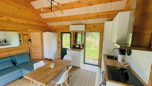 A wooden cabin interior with a dining table, chairs, a sofa, a kitchenette, and a large window. - Charming holiday home near the beach (Gedser)
