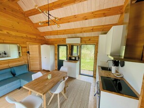 A wooden cabin interior with a dining table, chairs, a sofa, a kitchenette, and a large window. - Charming holiday home near the beach (Gedser)