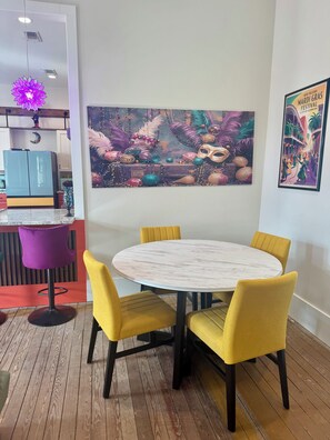 Ein Essbereich mit einem runden Tisch und gelben Stühlen, einem violetten Barhocker und einer Wand mit einem Gemälde zum Thema Mardi Gras.