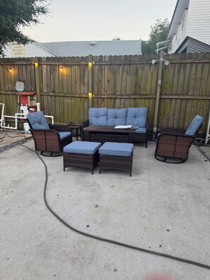 An outdoor patio area with a wooden fence, a blue cushioned sofa, wicker chairs, and a concrete floor.